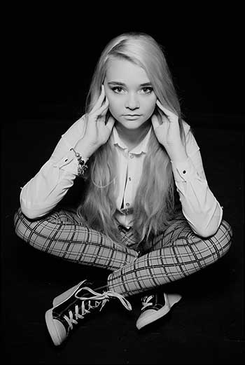 Black and white portrait photograph of a young woman sat cross legged in Rupert the bear chequered trousers.